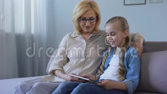长头发的孙女在平板电脑上看电影祖母在沙发上看电影视频的预览图