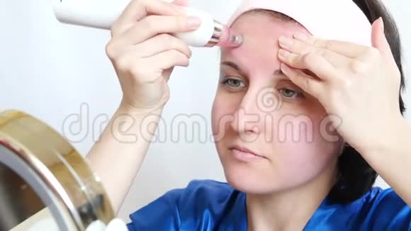 一个女孩带着一个装置在白色背景上做面部皮肤的微磨擦坐着镜子视频的预览图