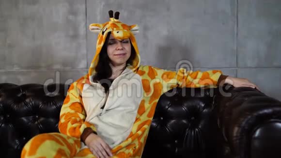 穿长颈鹿服装的女人坐在沙发上穿着长颈鹿滑稽睡衣坐在皮榻上微笑的年轻女子视频的预览图