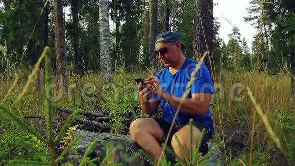 森林里一个戴着太阳镜和棒球帽的游客坐在一棵倒下的树上用电话交流视频的预览图