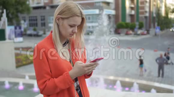 穿红色夹克的年轻漂亮女孩用手机站在喷泉背景下视频的预览图
