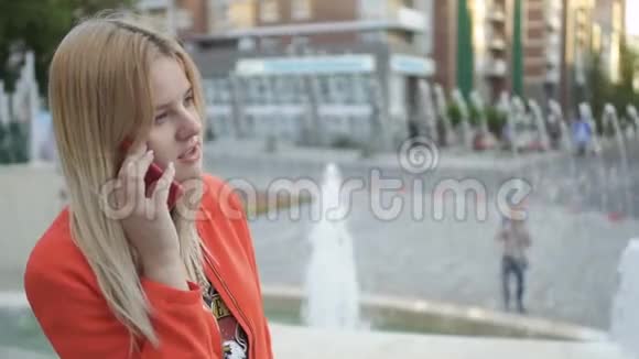 穿红色夹克的漂亮女孩站在喷泉背景下打电话视频的预览图