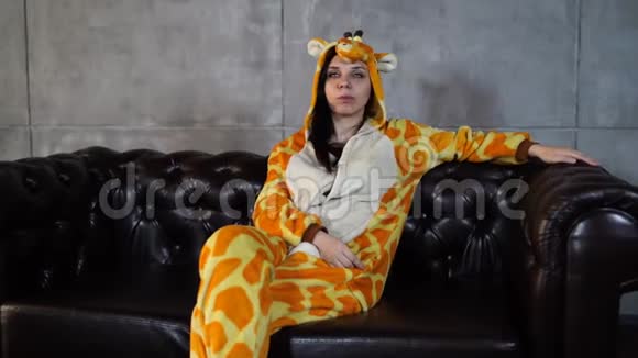 穿长颈鹿服装的女人坐在沙发上穿着长颈鹿滑稽睡衣坐在皮榻上微笑的年轻女子视频的预览图