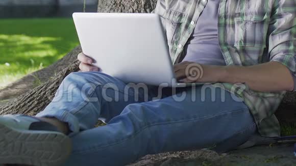 男性坐在树下在笔记本电脑上工作IT开发人员自由职业者视频的预览图