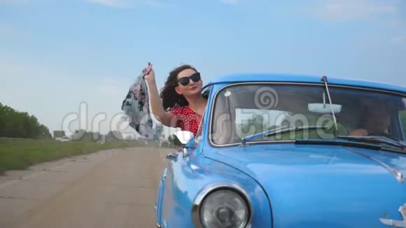 带着围巾的年轻女孩从老式车窗里探出来享受着旅行的乐趣女人看不出移动复古车视频的预览图