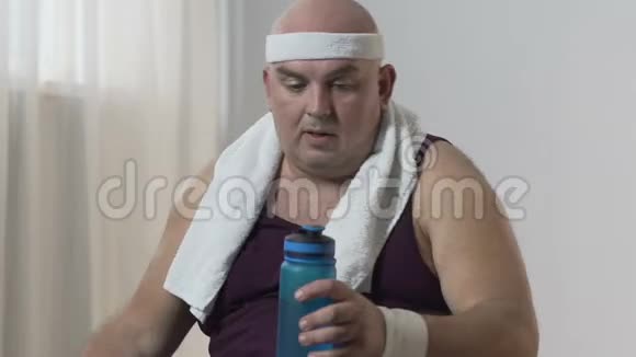 运动服的肥胖男性活动后坐在垫子上喝水视频的预览图
