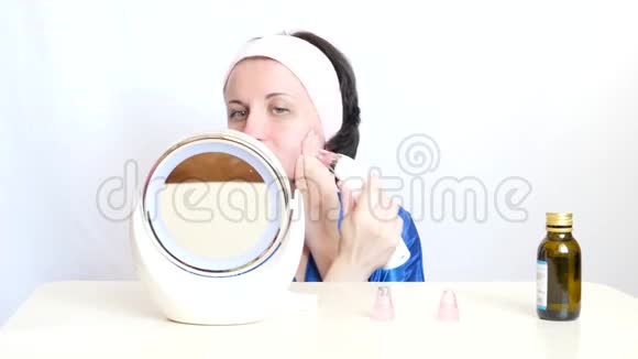 一个女孩带着一个装置在白色背景上做面部皮肤的微磨擦坐着镜子视频的预览图