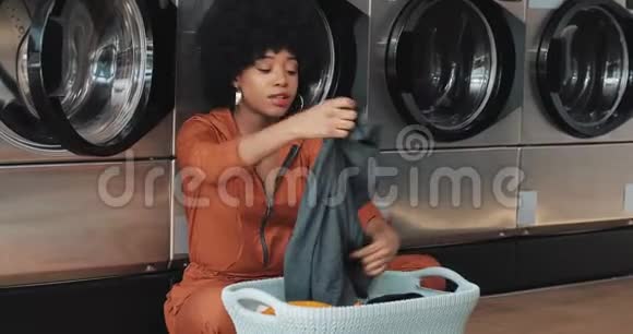 非洲裔美国年轻女性在洗衣前用篮子整理衣物然后坐在自助式公共场所洗衣服视频的预览图