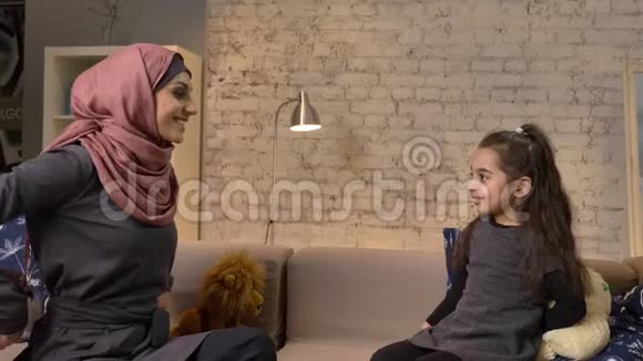 年轻的穆斯林母亲戴着头巾给她的女儿泰迪羊一家人坐在沙发上无所事事在家里安慰自己视频的预览图