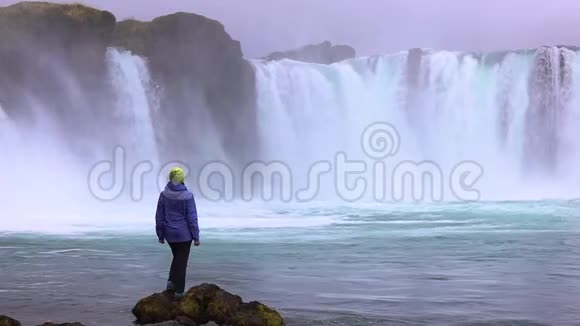 一个年轻的女人欣赏一个强大的狂暴瀑布沿着岩石边缘重重地落下岩石上落下一条清澈的小溪视频的预览图
