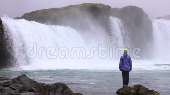 一个年轻的女人欣赏一个强大的狂暴瀑布沿着岩石边缘重重地落下岩石上落下一条清澈的小溪视频的预览图