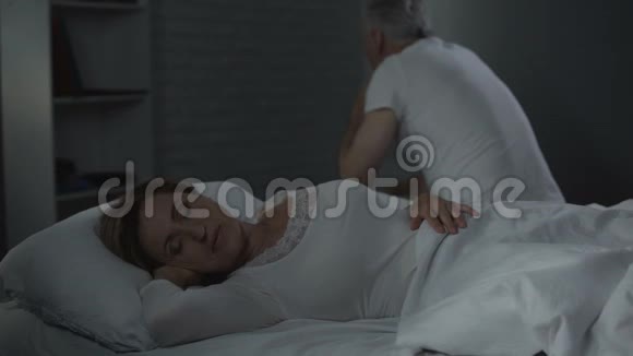退休男子坐在远远的床沿向后转身看着熟睡的女人视频的预览图