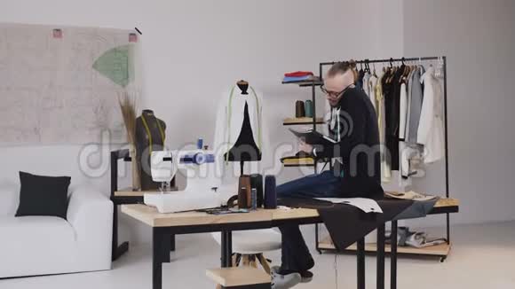 英俊的时装设计师坐在工作室的桌子上打电话他在笔记本上做笔记小胡子视频的预览图
