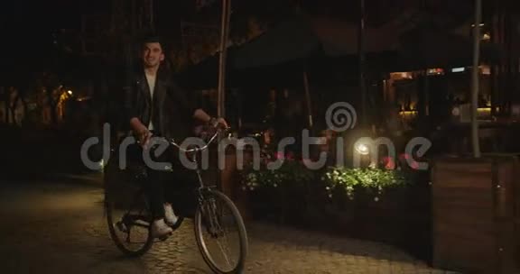 夜晚年轻人骑着自行车沿着可爱的老式咖啡馆的近景视频的预览图