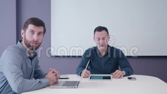 两个中年商人坐在会议室里桌上放着平板电脑和笔记本电脑礼貌地讨论视频的预览图