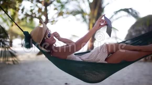 放松的女孩戴着太阳镜和帽子自拍手机销售手机躺在吊床之间的棕榈树1920x1080视频的预览图