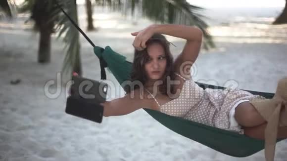 放松的漂亮女孩与手机销售手机自拍躺在吊床之间的棕榈树1920x1080视频的预览图