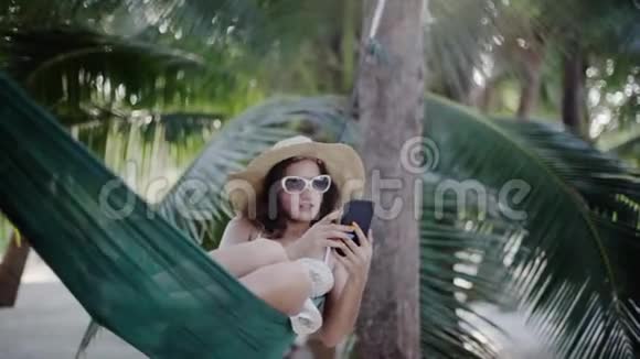 戴着太阳镜和帽子的放松的女孩用手机躺在棕榈树之间的吊床上1920x1080视频的预览图