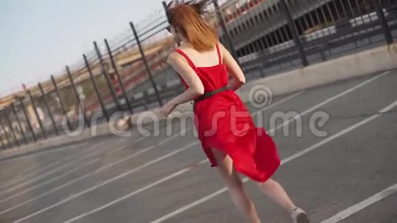 迷人的年轻红发女人穿着红色的裙子在街上奔跑在日落时玩得很开心慢动作视频的预览图