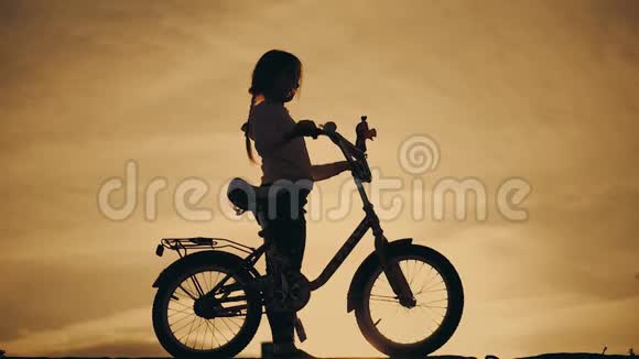 夕阳背景下一个女孩骑自行车的夏日风景剪影孩子在日落时骑自行车视频的预览图