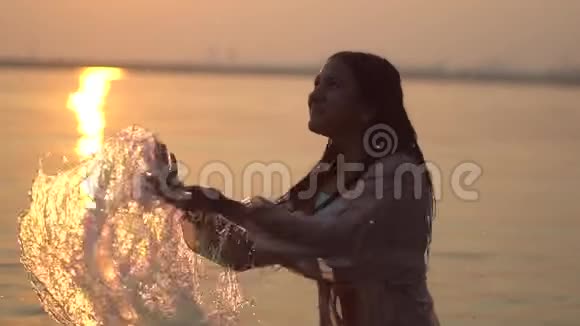 快乐的女孩一头乌黑的头发在夕阳下站在河里溅起双手慢动作关闭视频的预览图