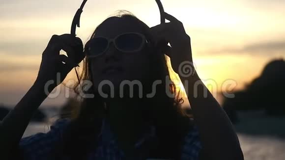 戴着耳机戴着太阳镜的年轻女子在令人惊叹的日落时分在海滩上慢动作地听音乐视频的预览图