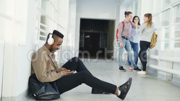 年轻英俊的非裔美国学生坐在白色走廊的地板上头上戴着耳机听音乐视频的预览图