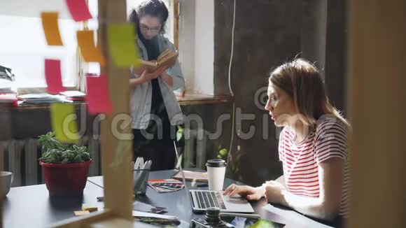 年轻的金发学生穿着休闲装忙着在笔记本电脑上打字而她的朋友正站在附近看书视频的预览图