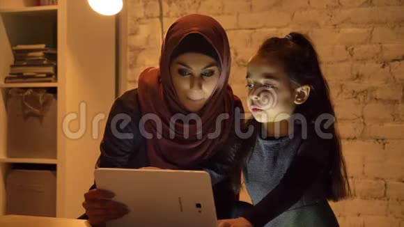 晚上一位戴着头巾的年轻母亲和她的小女儿坐在沙发上用平板电脑看一个有趣的节目视频的预览图