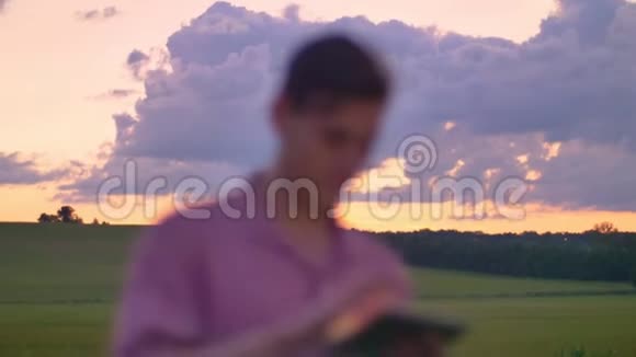 英俊的年轻人在石板上打字站在麦田或黑麦地上美丽的粉红色天空视频的预览图