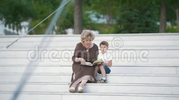 祖母给坐在公园楼梯上的孙子读了一本书视频的预览图