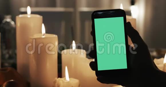 男性手拿着绿色屏幕和敲击的智能手机在背景中点燃蜡烛视频的预览图