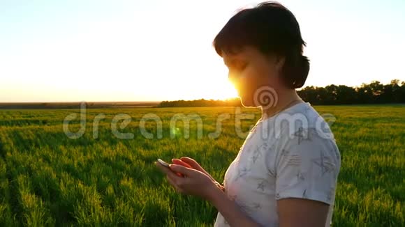 一个迷人的女孩正在绿色草坪上慢动作的夕阳背景下拿着一部智能手机视频的预览图