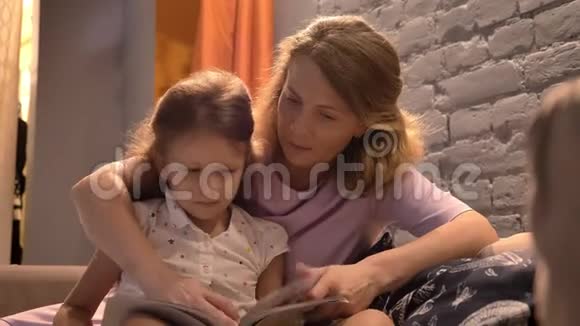 家长阅读书给小女孩妈妈和孩子坐在沙发上的现代客厅家庭观念室内视频的预览图