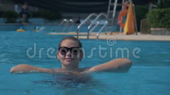 在阳光明媚的夏日戴太阳镜的欧洲女孩在外面的游泳池里溅起蓝色的水视频的预览图