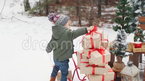 一个小男孩把礼物放在雪橇上概念圣诞快乐围巾和帽子红色的雪橇视频的预览图
