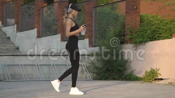 一个身材苗条的金发碧眼带着耳机在街上散步喝着水视频的预览图