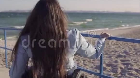 在春季或秋季坐轮椅的残疾青年妇女在靠近海洋的斜坡旁为残疾人移动视频的预览图