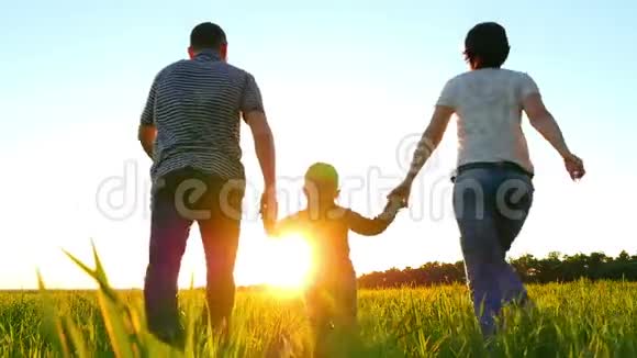 幸福的年轻家庭一个孩子在绿色的草坪上奔跑手牵着夕阳背景慢悠悠地走着视频的预览图