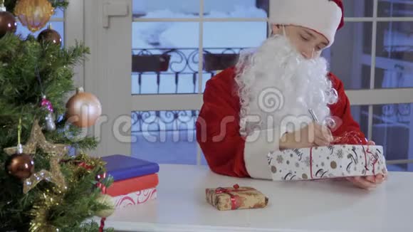 穿着圣诞老人服装的年轻人坐在桌子上在礼物上签名视频的预览图