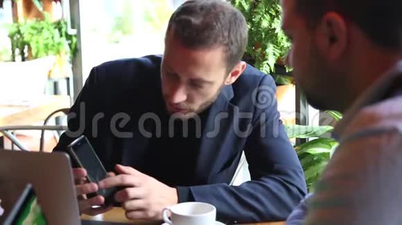 两个商人坐在咖啡馆的桌子上用笔记本电脑喝茶说话其中一个显示了另一个重要的信息视频的预览图