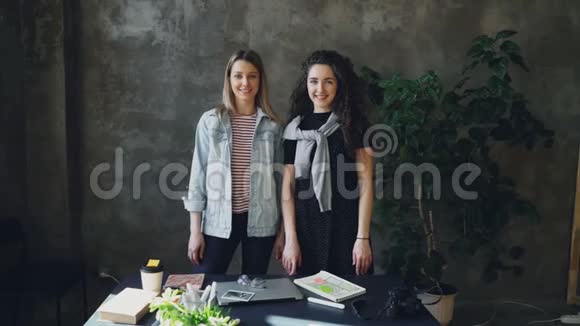两位年轻的女商务同事站在阁楼风格的办公室里摆姿势看着相机女孩视频的预览图