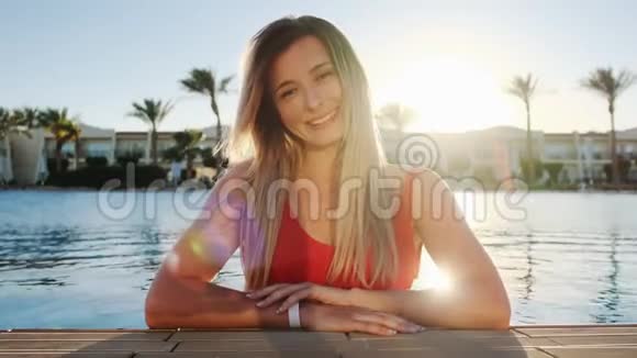 微笑的年轻女孩的肖像看着相机穿着红色泳衣的美女坐在蓝色游泳池里视频的预览图