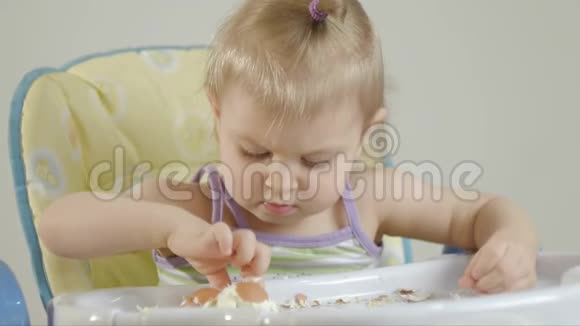 一个小女孩坐在一个儿童椅子上用一个煮熟的鸡蛋清理外壳并吃掉它视频的预览图