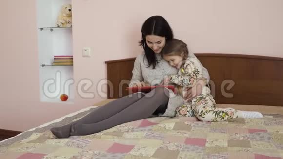 可爱的母亲和女儿使用平板电脑坐在家里的床上视频的预览图