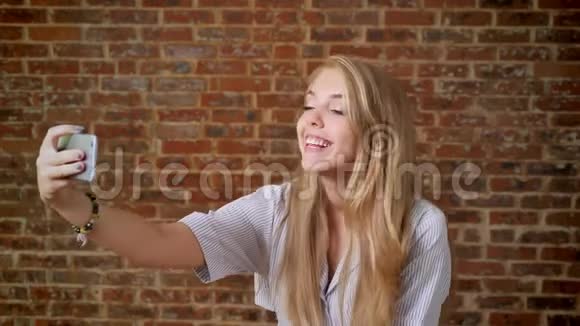年轻的白种人金发女郎自拍照片智能手机肖像背景砖墙视频的预览图