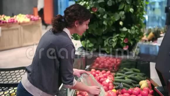 一个年轻漂亮的黑发女孩20正在水果和蔬菜过道里把西红柿放进塑料袋里视频的预览图
