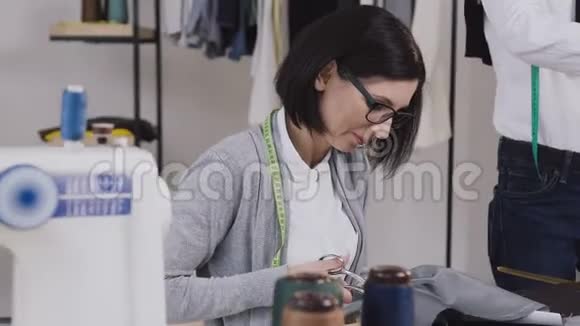 一个裁缝坐在桌子旁用剪刀剪灰色的材料用于缝小衣服视频的预览图