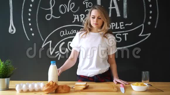 一个穿着睡衣的漂亮女人一件白色的t恤准备在厨房的家里吃早餐牛奶面包视频的预览图
