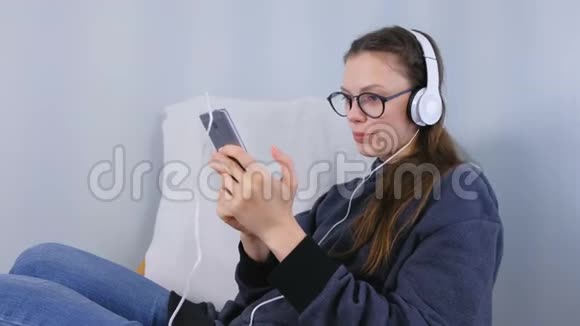 女人在智能手机上用耳机听音乐坐在扶手椅上唱一首歌视频的预览图